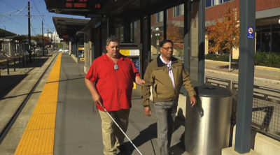Photo of a deafblind man with a cane walking with his SSP at the light rail station